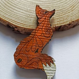 May include: A wooden fox-shaped brooch with intricate etched details. The fox is primarily orange-brown with a white tail tip. The brooch is set against a natural wood slice background.