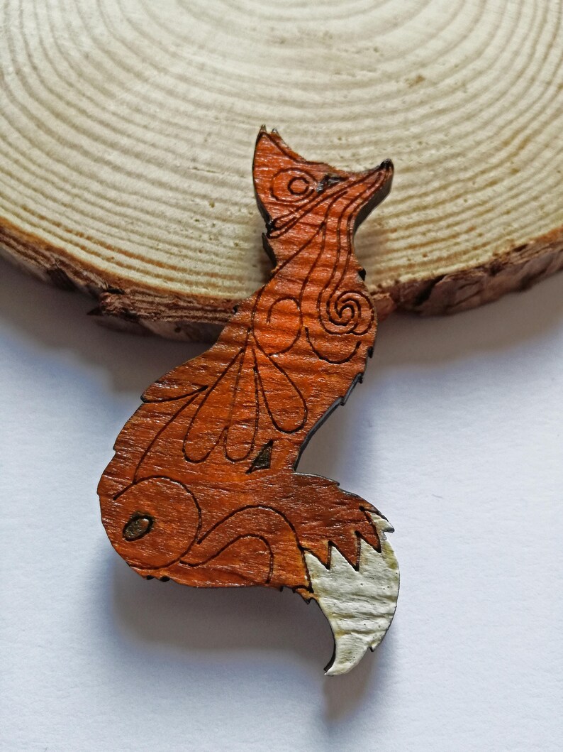May include: A wooden fox-shaped brooch with intricate etched details. The fox is primarily orange-brown with a white tail tip. The brooch is set against a natural wood slice background.
