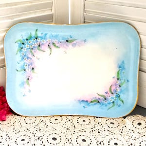 May include: A light blue porcelain tray with a gold rim and a hand-painted design of forget-me-not flowers.