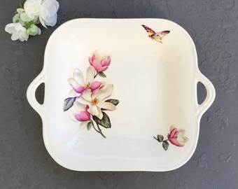 May include: White ceramic serving dish with two handles. The dish features a floral design with pink and white magnolia flowers and green leaves. A butterfly is painted in the upper right corner.