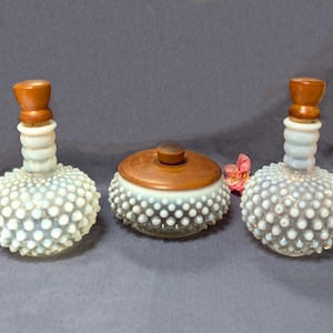 Pode incluir: Duas garrafas de vidro opalescente com textura de pontos e rolhas de madeira castanha, juntamente com um frasco de vidro redondo a condizer com tampa de madeira castanha. O vidro apresenta um padrão distinto de pontos brancos em relevo.