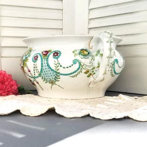 May include: A white ceramic planter with a floral design in shades of green, blue, and red. The planter has a large, rounded body and a single handle.