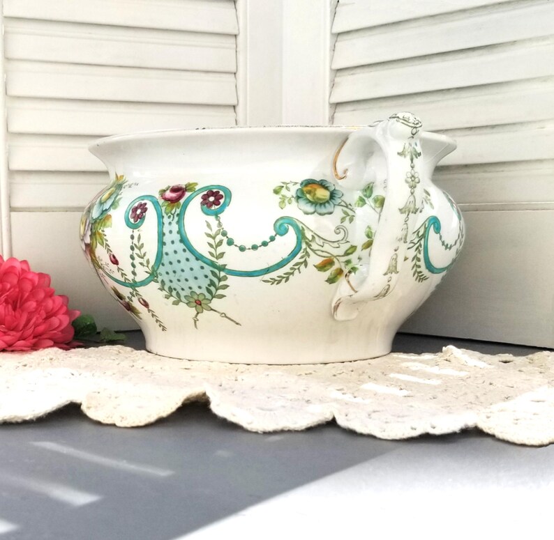 May include: A white ceramic planter with a floral design in shades of green, blue, and red. The planter has a large, rounded body and a single handle.