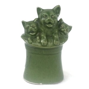 May include: A green ceramic figurine featuring three dogs. The dogs are positioned together, with one dog in the center and two on either side. The figurine has a cylindrical base and is painted in a solid green color.