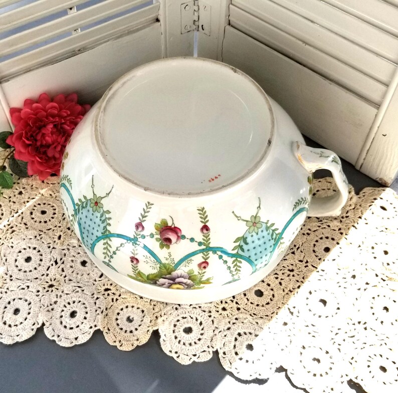May include: A white ceramic bowl with a blue and green floral design. The bowl has a handle on one side and is decorated with a repeating pattern of flowers and leaves.