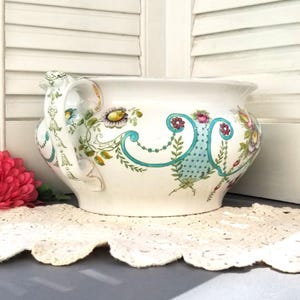 May include: A white ceramic planter with a floral design in shades of pink, blue, green, and yellow. The planter has two handles and a wide, round base.