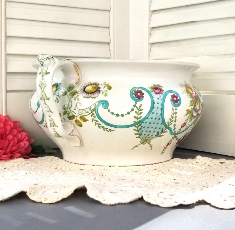 May include: A white ceramic planter with a floral design in shades of pink, blue, green, and yellow. The planter has two handles and a wide, round base.