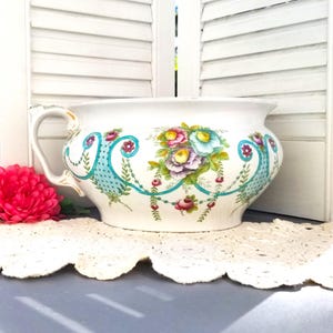 May include: A white ceramic planter with a floral design featuring blue, pink, and yellow flowers. The planter has a handle and a wide, rounded base.