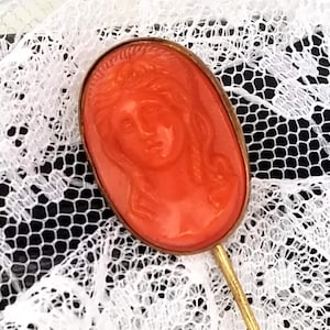 May include: A vintage orange cameo brooch with a woman's face in relief. The cameo is set in a gold-toned metal frame and attached to a long gold-toned pin.