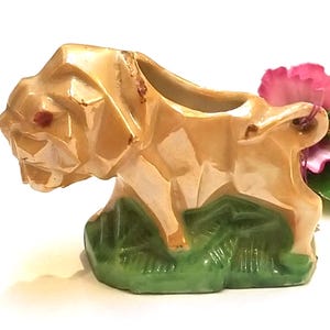 May include: A ceramic figurine of a dog with a faceted, light tan, pearlescent finish. The dog is depicted in a standing position on a textured, dark green base. The piece features a small opening on the back and has a pink flower attached to the tail.