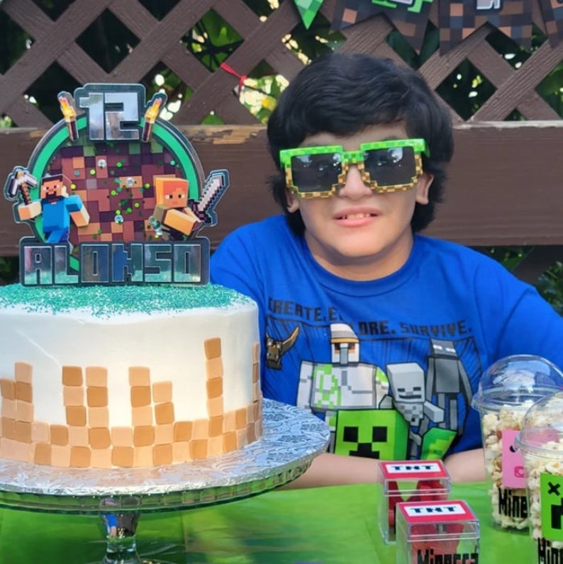May include: A Minecraft themed birthday cake with a green and brown frosting design. The cake is decorated with a topper that says "12 ALONSO" and features Minecraft characters. A boy wearing green Minecraft-themed glasses is sitting at a table with the cake. There are also Minecraft-themed party favors on the table.