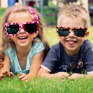 May include: Two children wearing colorful pixelated sunglasses. The girl is wearing pink and white sunglasses and a blue and white patterned shirt. The boy is wearing brown and orange sunglasses and a dark blue shirt. They are both smiling and lying on their stomachs in a grassy field.