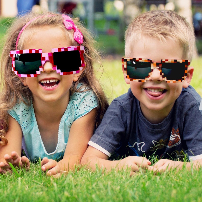 May include: Two children wearing colorful pixelated sunglasses. The girl is wearing pink and white sunglasses and a blue and white patterned shirt. The boy is wearing brown and orange sunglasses and a dark blue shirt. They are both smiling and lying on their stomachs in a grassy field.