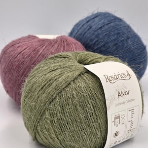 Könnte beinhalten: Drei Knäuel Garn in den Farbtönen Staubrosa, Olivgrün und Jeansblau. Das grüne Knäuel hat ein Etikett mit der Aufschrift "Rosários 4 Alvor Ecofriendly Collection". Das Garn scheint aus Naturfasern zu bestehen.