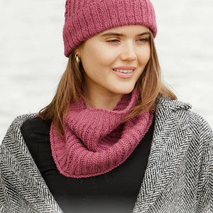 May include: A woman wearing a knitted hat and cowl in a deep rose color. The hat has a cable knit design, and the cowl is a simple ribbed knit. The woman is wearing a black top and a gray and white patterned coat. The image includes the text "DROPS DESIGN".