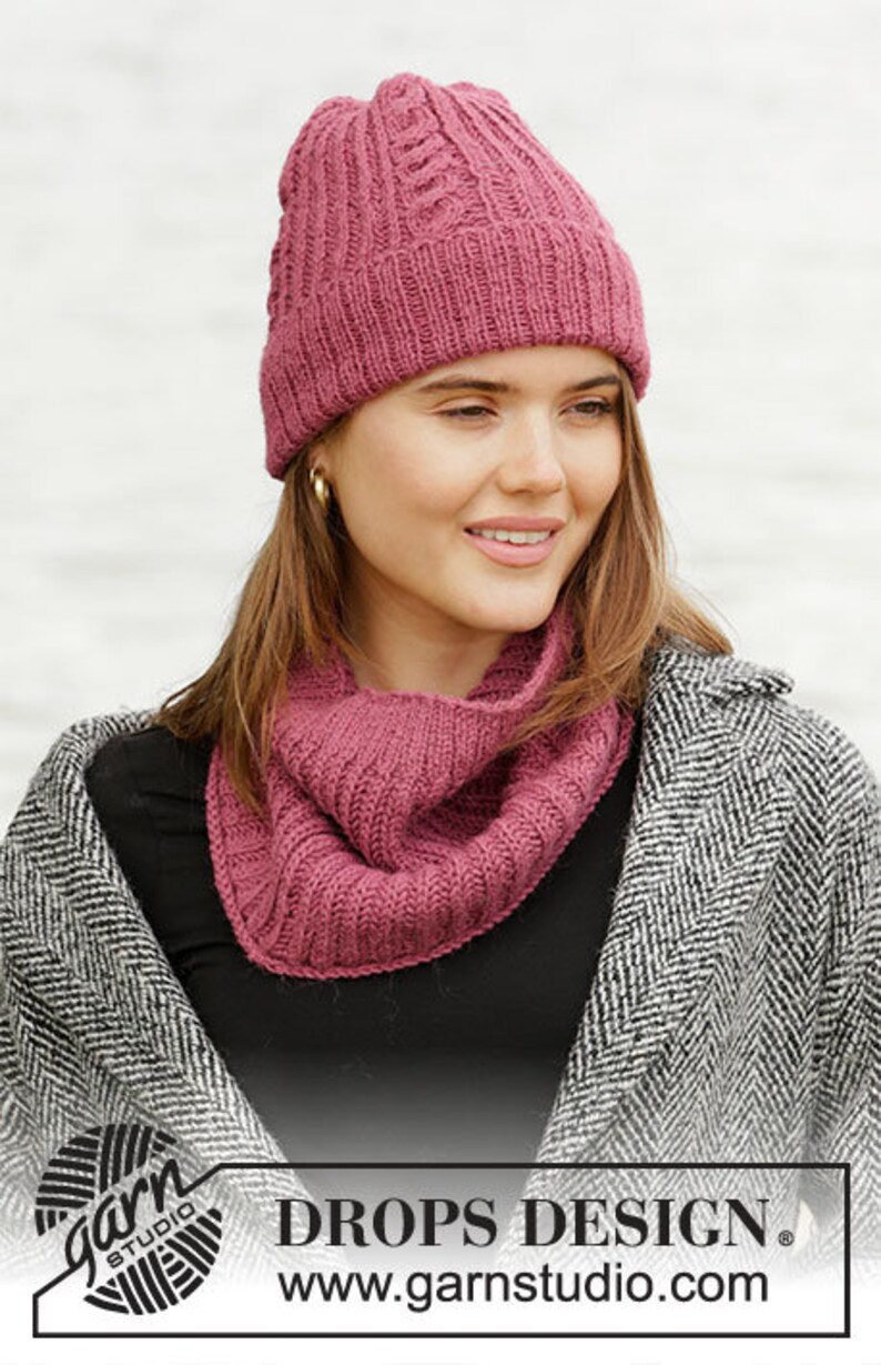 May include: A woman wearing a knitted hat and cowl in a deep rose color. The hat has a cable knit design, and the cowl is a simple ribbed knit. The woman is wearing a black top and a gray and white patterned coat. The image includes the text "DROPS DESIGN".