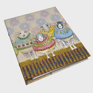 Op de afbeelding: Een notitieboek met een aquarel illustratie van vier schapen met kleurrijke truien. De schapen staan op een gebreide rand met een lichtbeige patroonachtergrond. De truien van de schapen zijn in verschillende kleuren, waaronder blauw, rood en geel.
