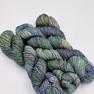 May include: A skein of yarn in shades of green, blue, and purple. The yarn is arranged in a loose coil.