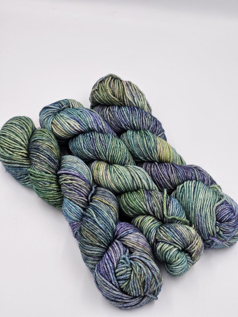 May include: A skein of yarn in shades of green, blue, and purple. The yarn is arranged in a loose coil.