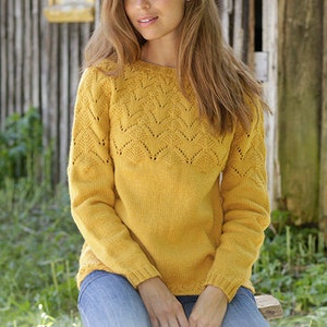 K&ouml;nnte beinhalten: Ein gelber Strickpullover mit einem zarten Spitzenmuster um den Ausschnitt und die Schultern. Der Pullover hat lange &Auml;rmel und eine lockere Passform. Das Model tr&auml;gt den Pullover mit einer blauen Jeans und sitzt auf einer Holzbank.