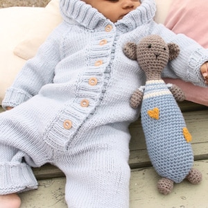 Puede incluir: Un pelele de punto azul claro con capucha y cierre de botones. El pelele lo lleva un beb&eacute; que sostiene un animal de peluche de ganchillo. El texto "DROPS DESIGN" y "www.GARNSTUDIO.com" es visible en la parte inferior de la imagen.