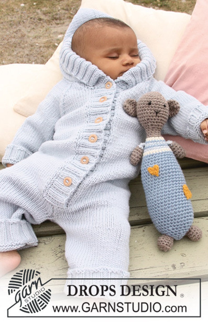 Puede incluir: Un pelele de punto azul claro con capucha y cierre de botones. El pelele lo lleva un beb&eacute; que sostiene un animal de peluche de ganchillo. El texto "DROPS DESIGN" y "www.GARNSTUDIO.com" es visible en la parte inferior de la imagen.