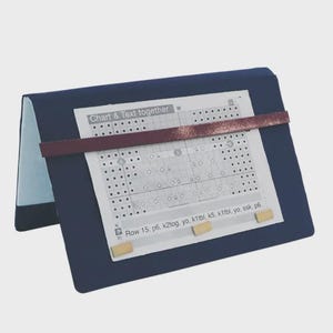 May include: A navy blue knitting chart holder with a maroon strap holding a knitting pattern in place. The text "Chart & Text together" is visible. Two small wooden magnets secure the pattern. The holder is open and angled.