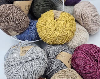 May include: A collection of multi-colored yarn balls in shades of yellow, purple, gray, brown, blue, and beige. Each ball features a paper label with product information, and several are arranged inside a white wire basket for storage.