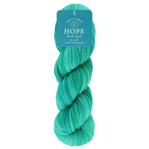 May include: A skein of hand-dyed yarn in shades of teal and green. The yarn is labeled "HOPE" by Simy's Studio. The tag is teal with white text and a small logo. The yarn is DK weight.