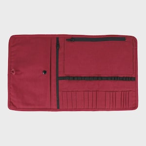 May include: A burgundy fabric art supply organiser, open and laid flat. It has a zippered pocket, a snap closure, and multiple small pockets for brushes or pens. Black zips and elastic bands provide contrast.