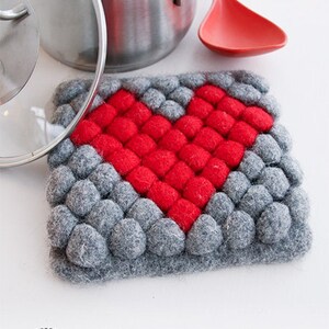 May include: A gray and red heart-shaped pot holder made of felt balls. The pot holder is perfect for protecting surfaces from hot pots and pans.