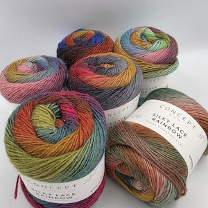 Puede incluir: Una colección de ovillos de lana coloridos. La lana presenta una mezcla de colores del arcoíris, incluyendo rosa, verde, naranja y azul. Cada ovillo está etiquetado con el texto "SILKY LACE RAINBOW". La lana es de seda merino extrafina.