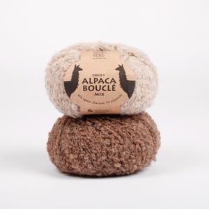 May include: Two stacked balls of textured yarn. The top ball is light beige and the bottom ball is medium brown. A tan paper label wraps around the top ball, featuring the text DROPS ALPACA BOUCLÉ MIX and illustrations of two alpaca silhouettes.