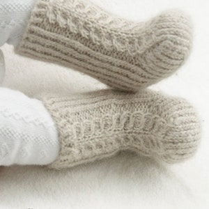 May include: Close-up of two light beige knitted socks with a cable knit pattern. The socks are made of a soft, textured yarn and are designed for warmth and comfort. The socks are a neutral color, suitable for various outfits.