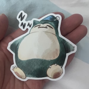 K&ouml;nnte beinhalten: Ein Aufkleber mit einer Aquarellillustration eines schlafenden Snorlax-Charakters. Das Snorlax ist in Gr&uuml;n-, Wei&szlig;- und Braunt&ouml;nen gehalten und tr&auml;gt eine blaue M&uuml;tze. Der Aufkleber hat einen wei&szlig;en Rand und den Text "Zzz".