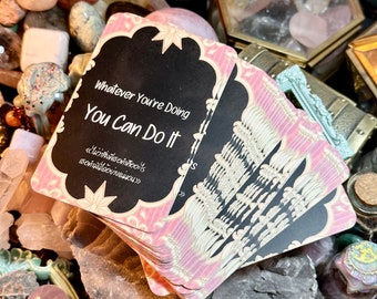 Könnte beinhalten: Ein Stapel rosa und schwarzer Affirmationskarten mit dem Text "Whatever You're Doing You Can Do It" auf der obersten Karte. Die Karten sind von verschiedenen Kristallen und Steinen umgeben.