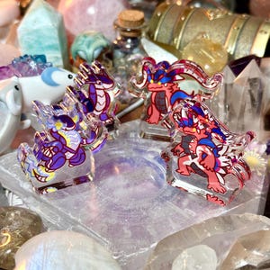 Puede incluir: Cuatro figuras de dragones de acrílico en tonos morado, rojo y azul. Los dragones están sobre bases transparentes, con cristales y otros objetos decorativos en el fondo. Las figuras tienen un diseño de dibujos animados.