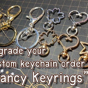 Puede incluir: Surtido de llaveros de oro y plata con diferentes formas, incluyendo un corazón, una estrella y un trébol. Los llaveros están sobre una superficie gris texturizada. "Upgrade your custom keychain order" y "Fancy Keyrings" están escritos en texto marrón sobre la superficie.