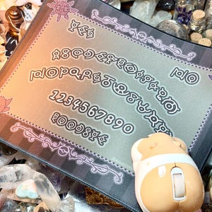 May include: A decorative mousepad with a gradient background, featuring a stylized alphabet, numbers, and symbols. The border has a floral design. A beige and white mouse shaped like a hamster is on the right side of the pad.