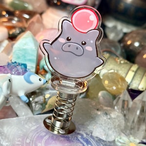 Puede incluir: Un juguete de escritorio de acrílico transparente con forma de cerdo con una burbuja rosa en la parte superior, montado sobre una base de resorte plateada. El cerdo tiene un cuerpo gris con una cara linda. Rodeado de cristales y otros objetos decorativos.