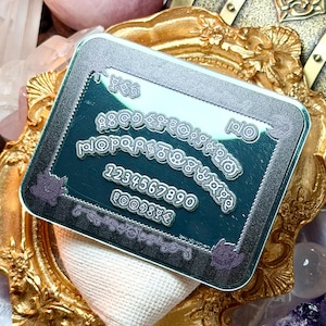May include: A teal and silver Ouija board with a decorative border and the alphabet, numbers, and the words "yes" and "no". The board is set on a gold ornate frame with a white cloth.