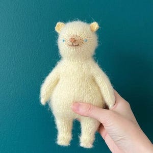 May include: A small, pale yellow knitted bear toy with a fuzzy texture. The bear features two tiny blue eyes, a small brown stitched nose, and a simple stitched mouth. The ears are rounded and the body has a soft, rounded shape with short limbs.
