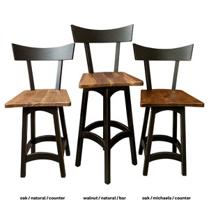 May include: Three wooden bar stools with black metal frames. The stools have a curved back and a wide, flat seat. The stools are made of oak and walnut wood. The stools are shown in three different finishes: natural, natural, and Michaels.