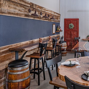May include: A rustic wooden bar with black metal stools and a wooden countertop. The bar has a dark blue chalkboard behind it and a red door in the background. There are several wine bottles on a shelf above the bar.