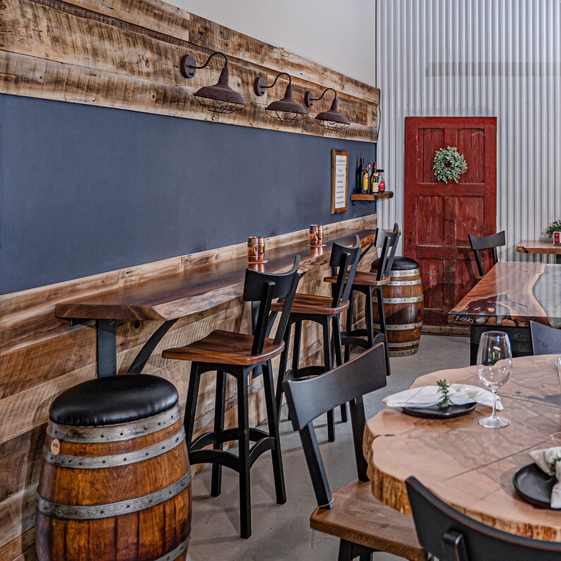 May include: A rustic wooden bar with black metal stools and a wooden countertop. The bar has a dark blue chalkboard behind it and a red door in the background. There are several wine bottles on a shelf above the bar.