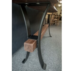 May include: Close-up of a wooden table leg with a black metal base. The leg is made of solid wood and has a natural finish. The metal base is curved and has a black powder coat finish.