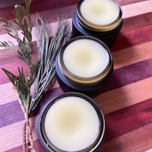 Op de afbeelding: Drie zwarte potten met gouden deksels gevuld met een lichtgele balsem of crème. De potten staan op een gestreept houten oppervlak.