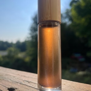 May include: A clear glass roll-on bottle filled with a bronze-colored liquid. The bottle has a wooden cap. The background is blurred, showing a wooden surface and green foliage. The bottle is standing upright in the sunlight.