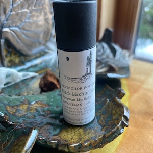 May include: A black tube of lip balm with a white label that reads "PETRICHOR POTION Black Birch and Apricot Beeswax Lip Balm 100% VEGAN 1 oz". The label also features a small illustration of three trees.