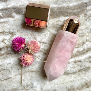 May include: A rose quartz facial roller with a gold-coloured cap and a small, gold-coloured square container. The roller is next to a small arrangement of dried pink flowers. The objects are on a marble surface.